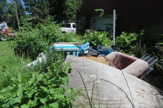 Before: A pile of old furniture, boards and junk in weeds beside a red building